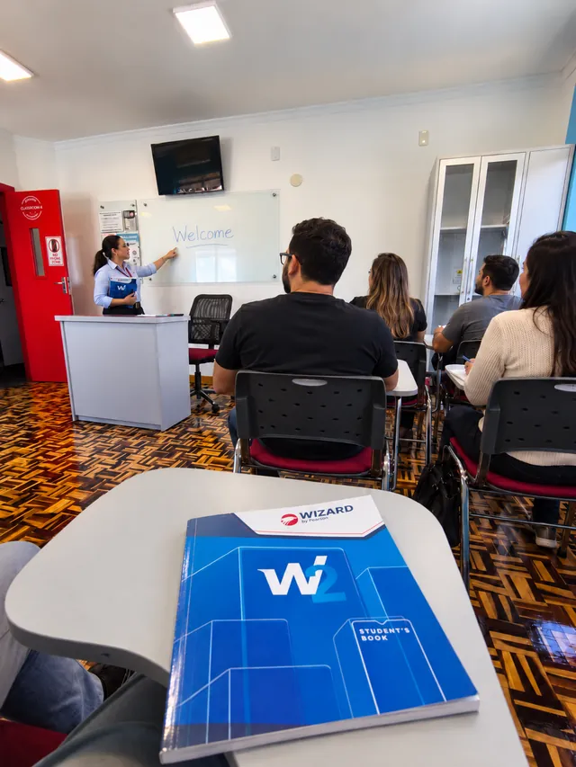 Aula presencial de adultos: a professora escreve Welcome no quadro e, em primeiro plano, o livro W2 da Wizard sobre a carteira