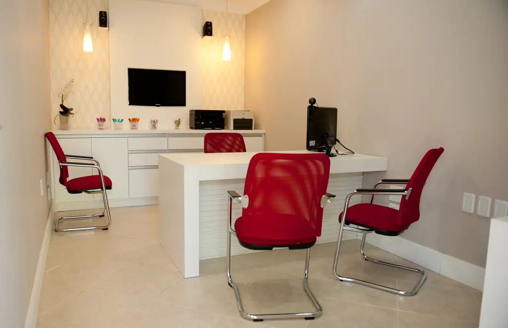 Sala de atendimento com mesa branca, cadeiras vermelhas e televisão