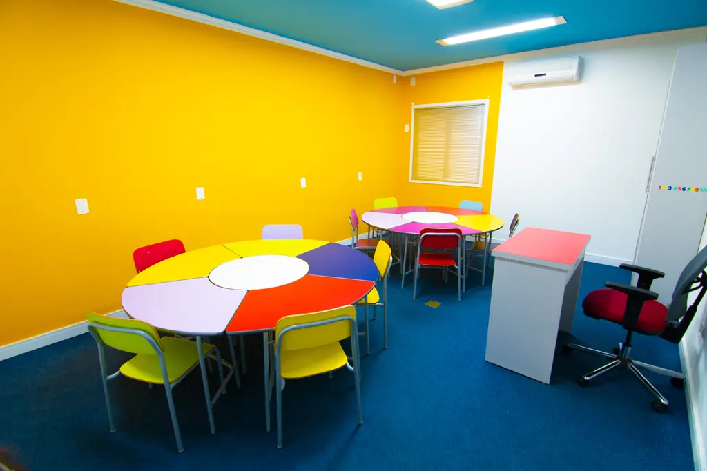 Sala de aula infantil com parede amarela, mesa redonda colorida e carpete azul